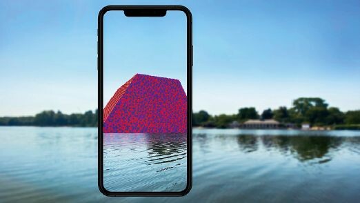 Landschaft mit See, vor der ein Smartphonerahmen zu sehen ist, in dem eine monumentale, angeschnittene Skulptur auf den See projiziert erscheint.