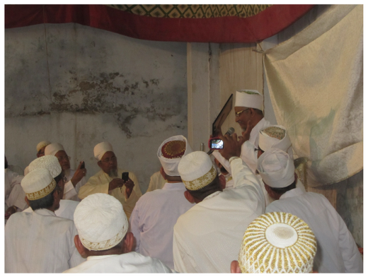 Figure 13.9: Above: picture taken from afar in the musāfir khāna (pilgrim lodge) in Ahmedabad before the Dāʿī reveals a naṣṣ scroll to a crowd of believers. Below: The Dāʿī reveals a framed copy of a fragment of a naṣṣ scroll during the ʿurs ceremony in Ahmedabad, Gujarat, while believers take pictures with their smartphones.