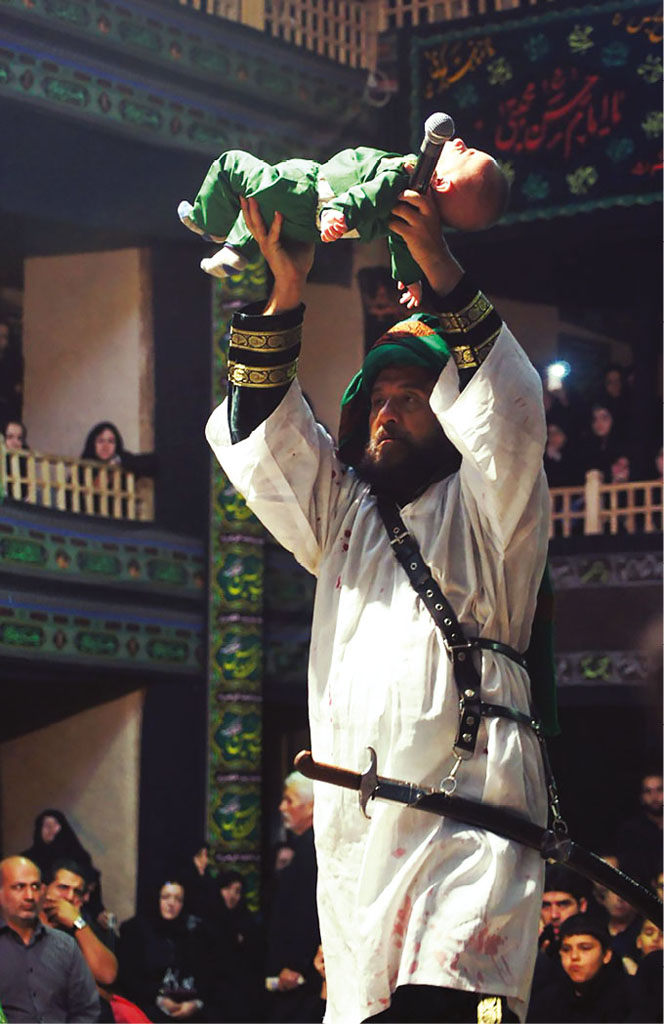 A bearded man wearing a green turban and white tunic lifts a baby into the air