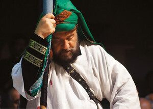 A bearded man wearing a green turban and white tunic