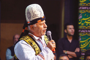 A man wearing a domed felt hat and embroidered waistcoat holds a microphone