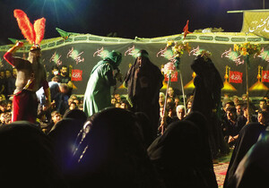 A man wearing a red feathered helmet wields a chain whip over a group of black and green clad figures