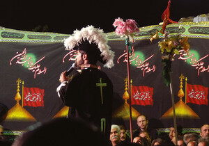 A man wearing a cocked hat and a jacket with a Christian cross on the back holds a microphone