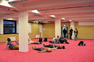 Circa 12 Menschen liegen oder sitzen auf einem pinken Teppich in einem Ausstellungsraum. Drei stehende Menschen betrachten sie.