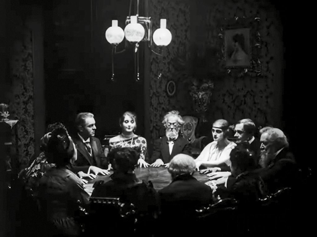 3.1 Eine Abendgesellschaft, darunter Dr. Mabuse und das Paar Told, sitzt, mit den Händen flach auf den Tisch gelegt, in einer Runde.
