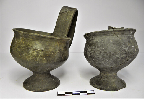 Footed cups with vertical handles connected to the rim. The left cup (NAM P4696) has a marked difference in height. Without its handle, it varies between 7.6â9.8 cm due to the fact that the whole vessel is skewed. The right cup (NAM P4697) has the same skewness, height 8.1â9.1 cm. Traces of the potterâs wheel visible under the foot, with clear wheel marks on the interior.